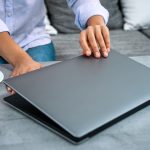 Widok dłoni kobiety otwierającej pokrywę laptopa. W pobliżu jest biała filiżanka kawy.