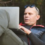 Zbliżenie: mężczyzna turysta leżący na plecach, opierając się na plecaku, z laptopem, pisanie na maszynie, blogowanie, przeglądanie w lesie