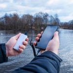 Mężczyzna ładuje smartfon powerbankiem. Telefon w dłoni ładuje się przenośną ładowarką na tle jeziora.