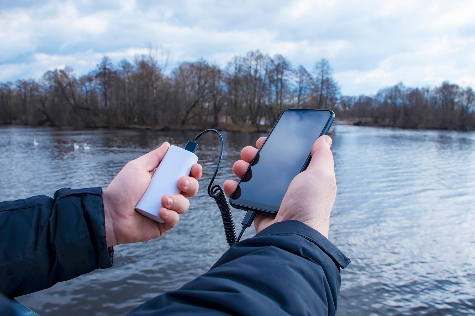 Mężczyzna ładuje smartfon powerbankiem. Telefon w dłoni ładuje się przenośną ładowarką na tle jeziora.
