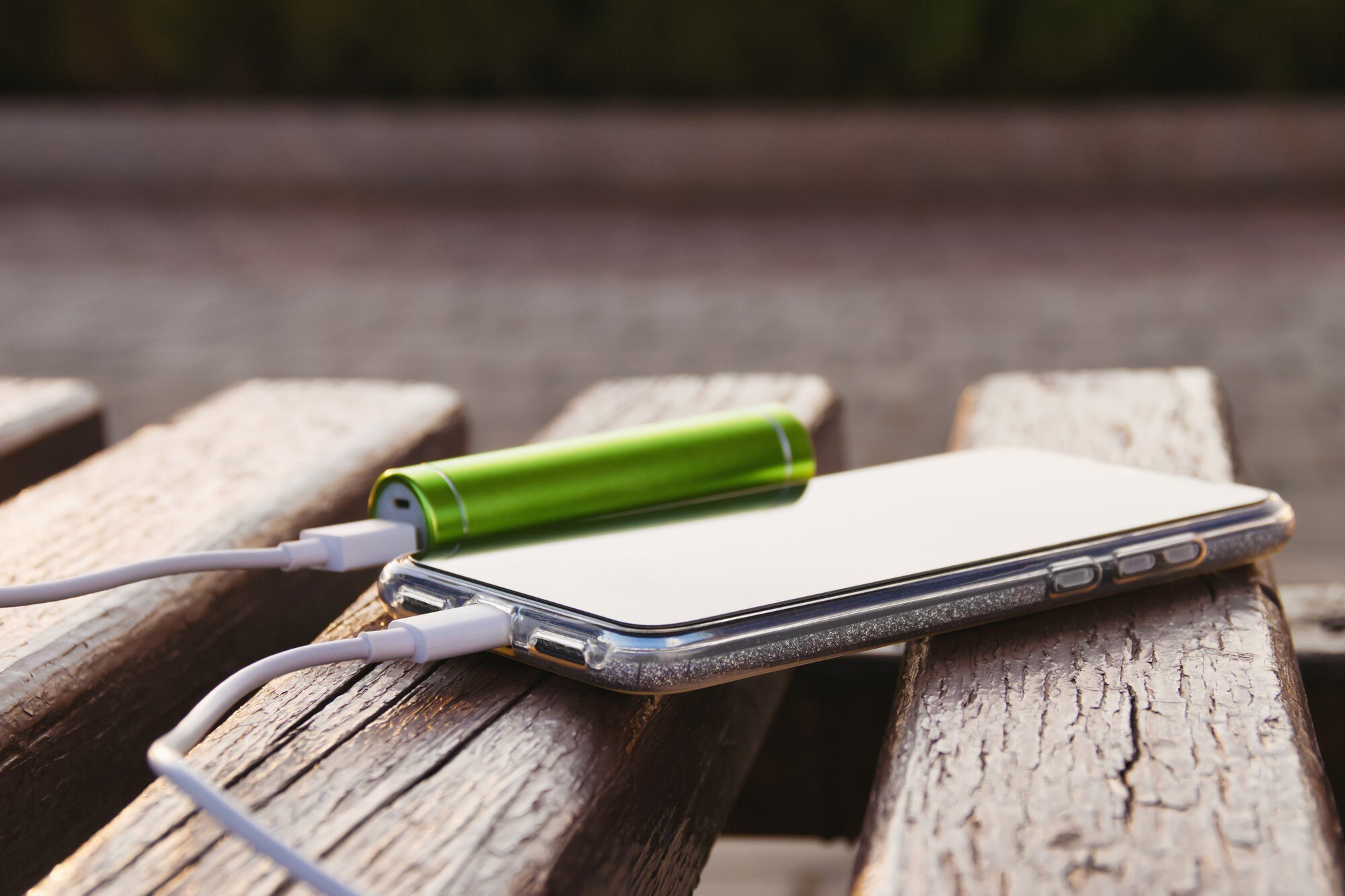 Smartfon i zewnętrzny power bank leżą na drewnianej ławce w parku podczas ładowania