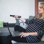 Widok z boku kobiety w ciąży z laptopa za pomocą pilota zdalnego sterowania
