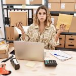 Młoda latynoska kobieta trzymająca paczkę i kopertę siedzi na biurku, nadymając policzki z zabawną twarzą. usta napompowane powietrzem, łapiąc powietrze.