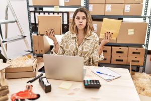 Młoda latynoska kobieta trzymająca paczkę i kopertę siedzi na biurku, nadymając policzki z zabawną twarzą. usta napompowane powietrzem, łapiąc powietrze.