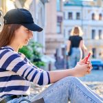Młoda nastolatka w miejskim stylu siedząca na chodniku z smartfonem