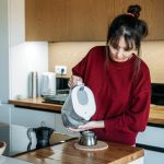 Person pours filtered water from pitcher into base of moka pot on wooden counter manual espresso