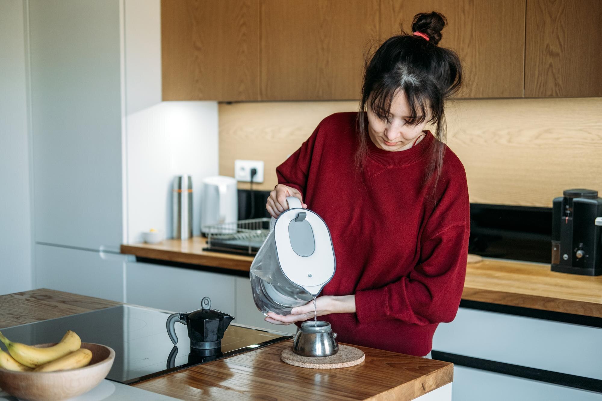 Person pours filtered water from pitcher into base of moka pot on wooden counter manual espresso