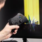 A frustrated gamer39s hand holds a black controller aggressively positioned against a broken computer monitor with a shattered screen and bright pixel lines