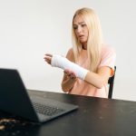 Portret smutnej rannej młodej kobiety ze złamaną ręką owiniętą w bandaż gipsowy po konsultacji wideo online za pomocą laptopa