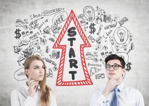 Portret biznesmena w okularach i jego blond kolega stojący przy betonowej ścianie z narysowanym na nim szkicem startowym.